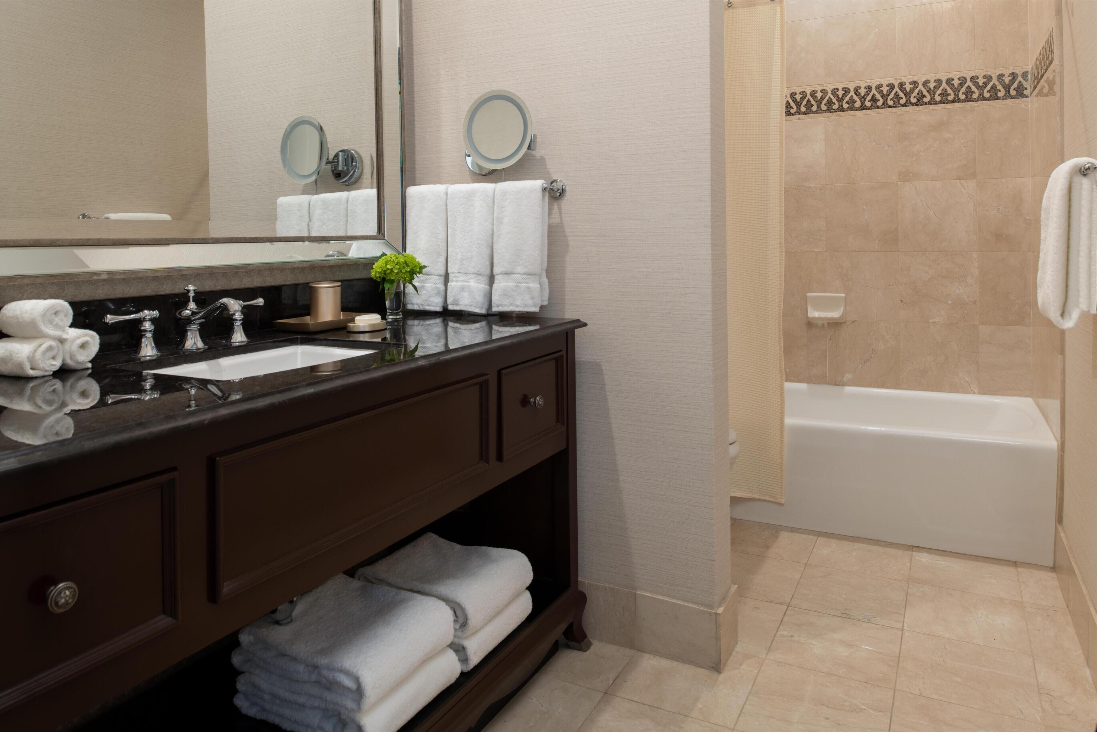 Un baño moderno con un tocador de madera oscura, dos lavamanos, un espejo grande, un espejo redondo, una bañera blanca y una zona de ducha.