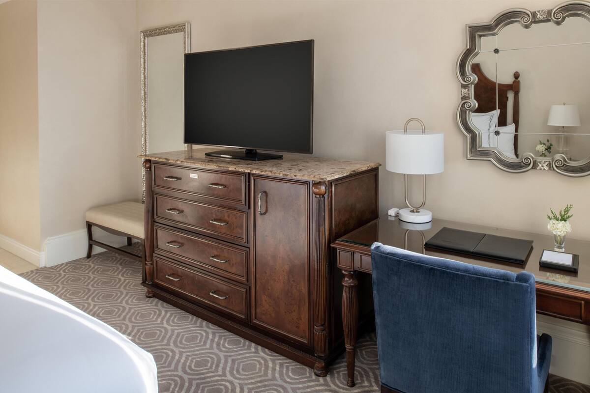 Een hotelkamer met een houten dressoir, een televisie, een bureau met een lamp, een stoel en een grote spiegel.