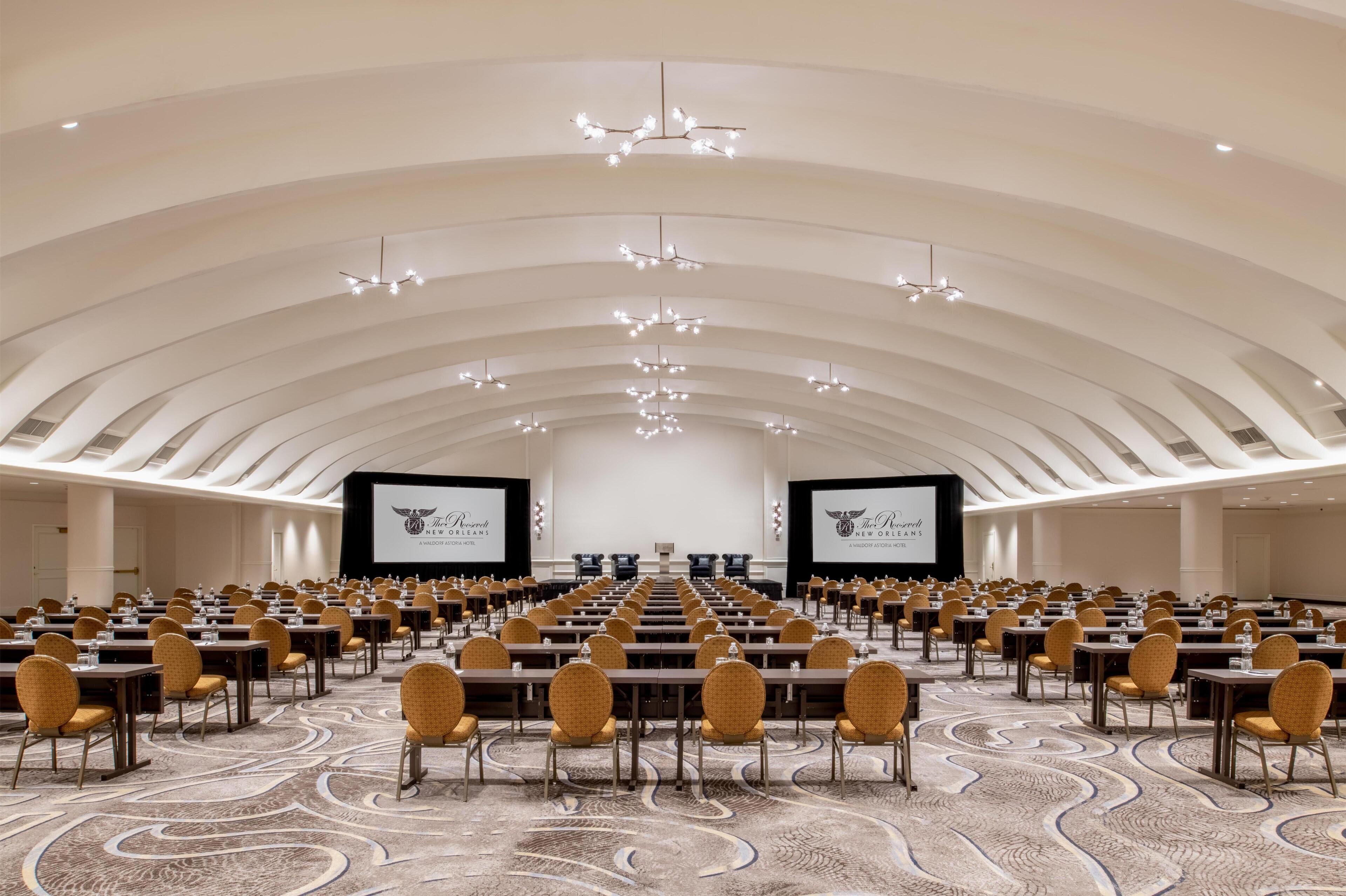 A large conference room with rows of tables and chairs, a high ceiling with arched design, and two large screens at the front.