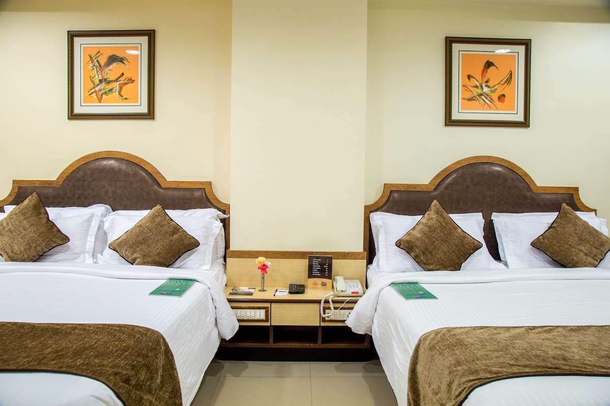 A hotel room with two beds, each with white linens and brown headboards. There are framed pictures on the walls, a bedside table with a phone and a lamp, and a small card on the bed.