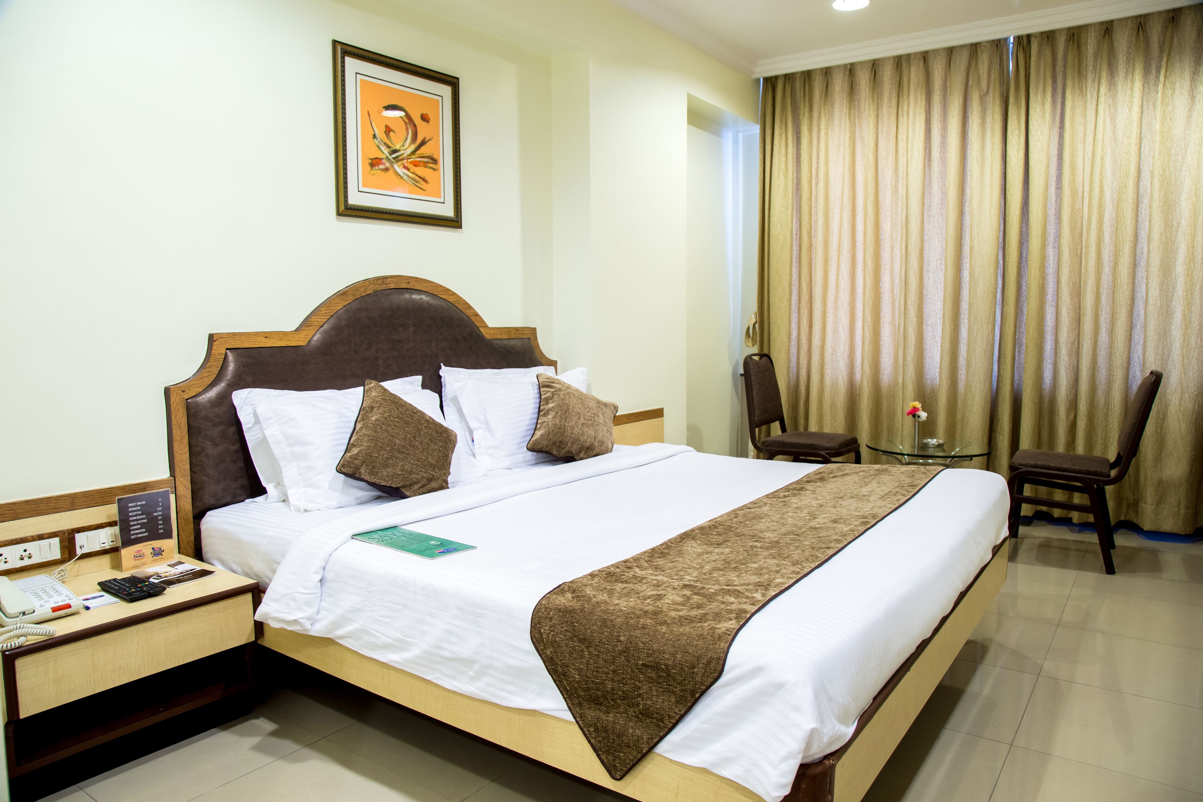 A hotel room with a large bed, a bedside table with a phone and a notepad, a small table with a flower, and a chair.