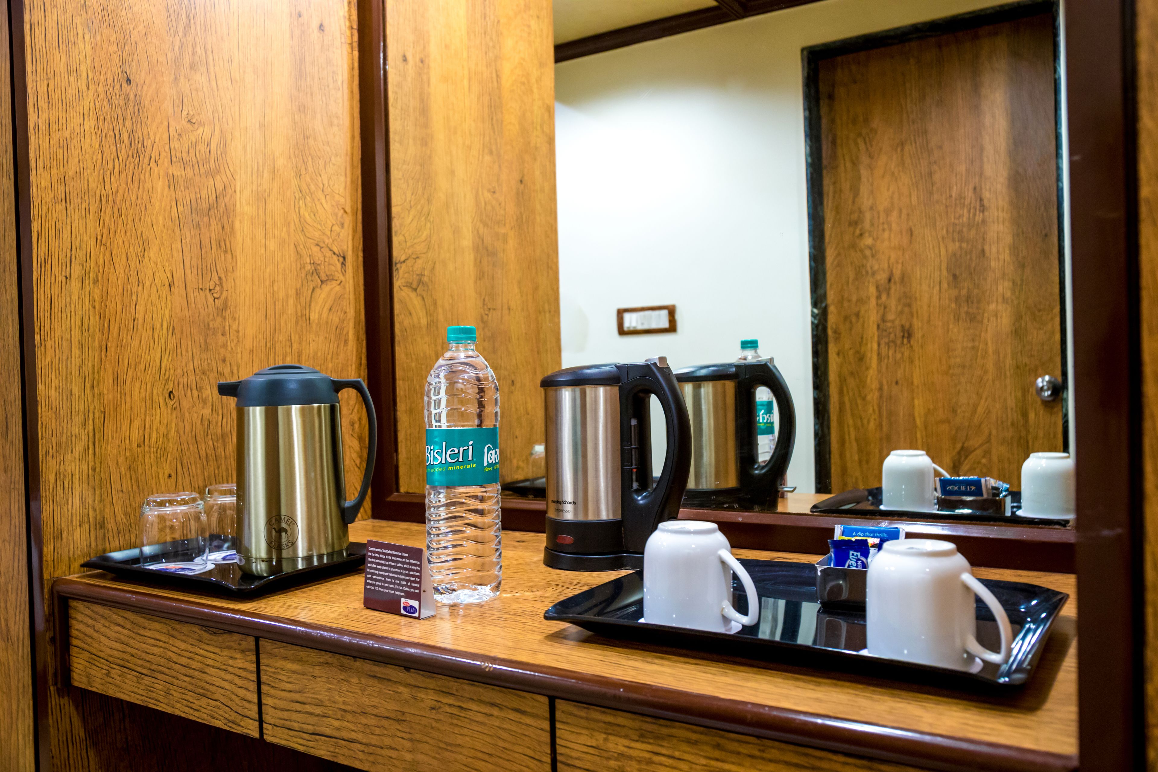 Zona de desayuno en una habitación de hotel, con mostrador de madera, cafetera, botella de agua y bandeja con tazas y una taza.