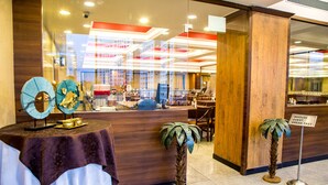 Ein Hotel-Empfangsbereich mit Holztresen, Schild für Frühstück im Haus und zwei dekorative Zimmerpflanzen.