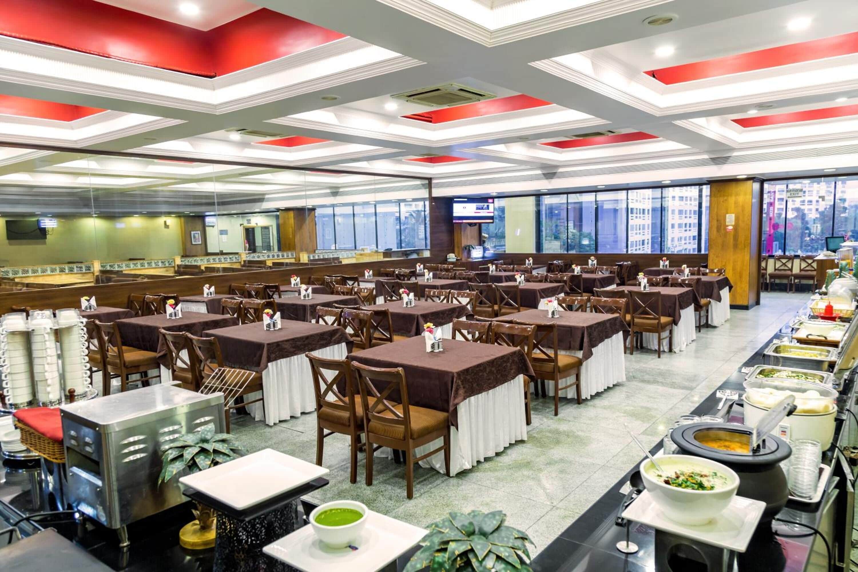 A large dining hall with multiple tables and chairs, a buffet line with various food items, and a television screen on the wall.