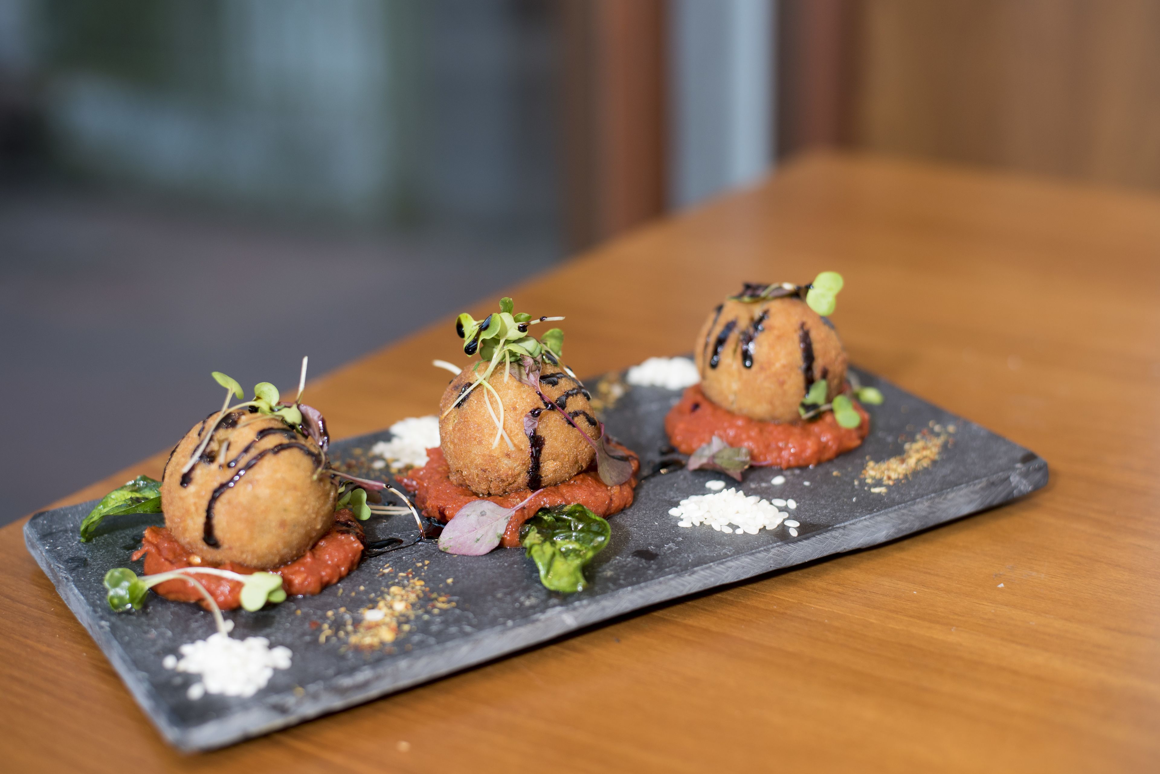 A slate serving board with three round, breaded items topped with greens and sauce, placed on a wooden table.