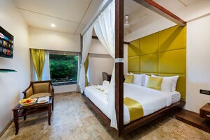 Una cama con dosel y cuatro columnas, una silla de madera con cojín, una mesita con cesta de fruta y una ventana con cortinas.
