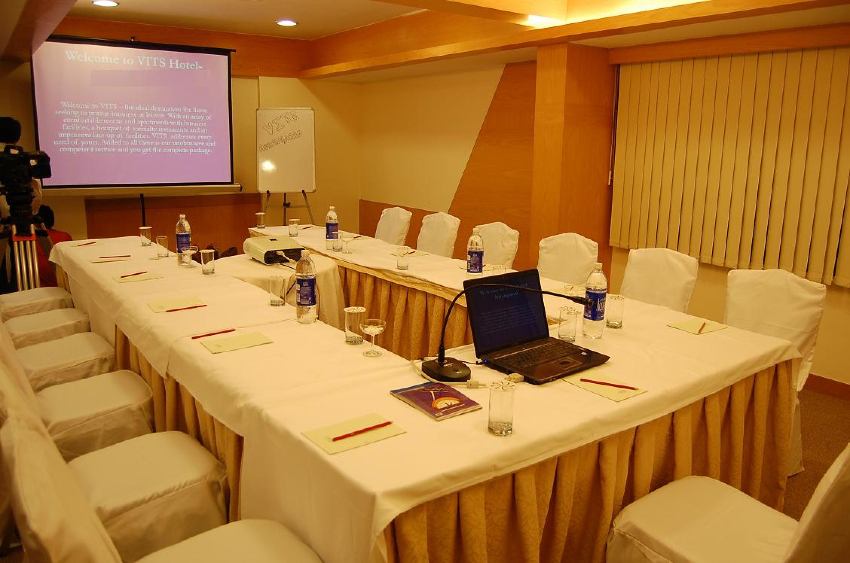 A conference room with a long table, chairs, a laptop, and a projector screen displaying a welcome message.