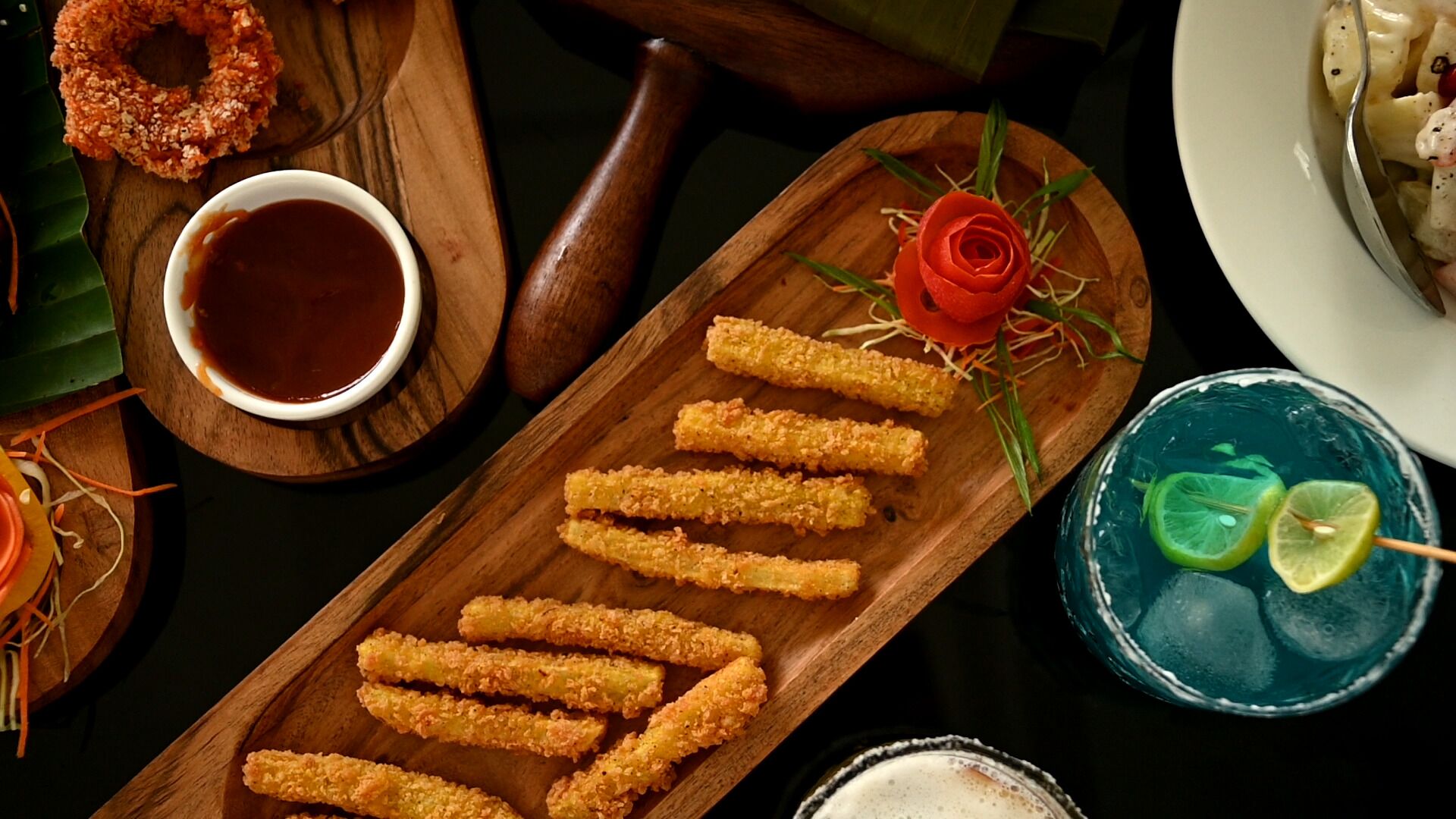 A wooden serving board with breaded items, a small bowl of sauce, a rose, and a cocktail with lime slices.