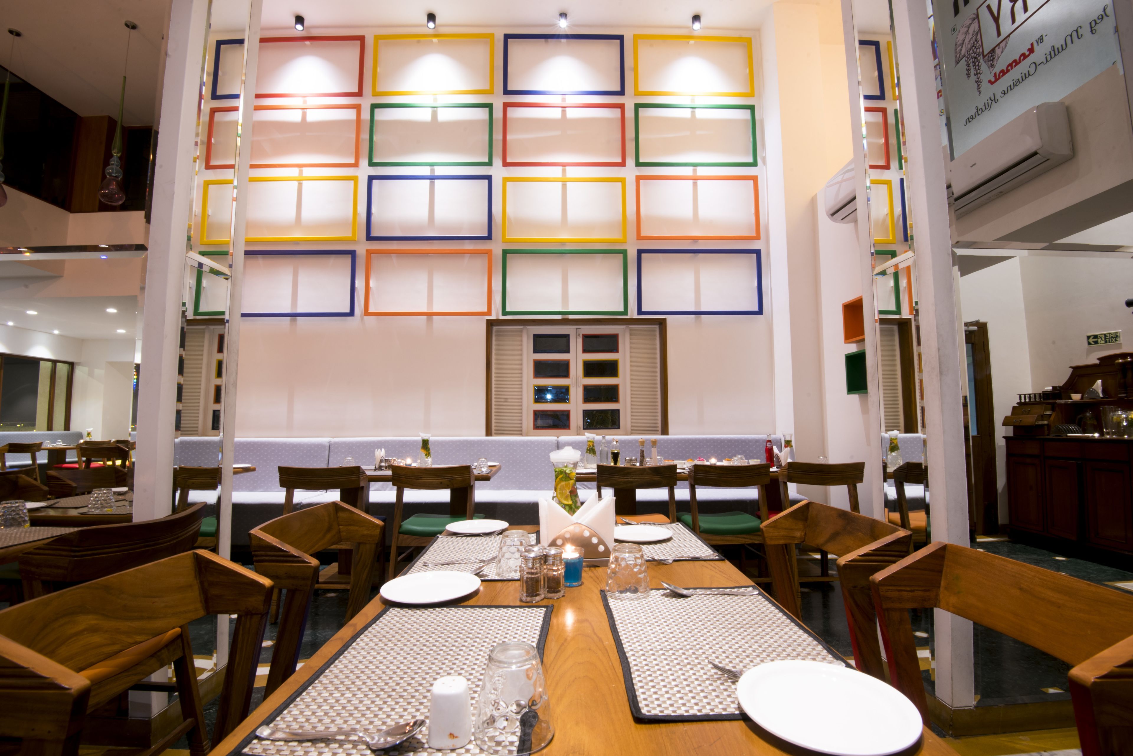A dining area with wooden tables and chairs, a central decorative vase, and a wall with colorful geometric patterns.