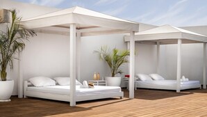 Une chambre à coucher moderne en plein air, avec deux lits blancs sous des baldaquins blancs, un plancher en bois et des plantes en pot.