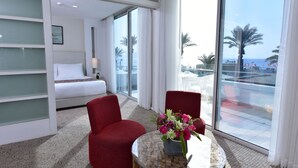 Une chambre d’hôtel avec un lit, un fauteuil rouge, une petite table sur laquelle est posé un vase de fleurs, et une grande fenêtre donnant sur des palmiers et la mer.
