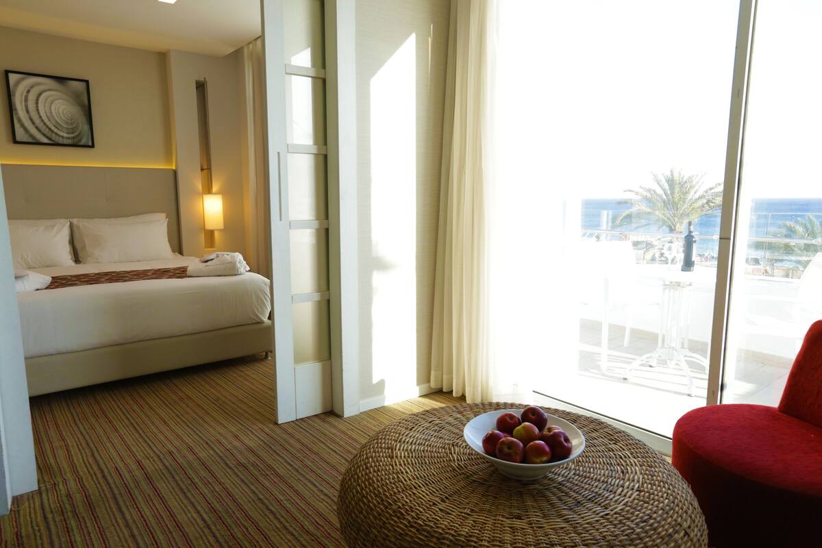 Une chambre d’hôtel avec un lit, un panier de fruits, un fauteuil rouge et une vue sur la ville grâce à de grandes fenêtres.