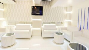 Une pièce moderne aux couleurs claires, avec un téléviseur à écran plat fixé au mur, deux canapés blancs et une rangée de lampes verticales bleues sur le mur de droite.