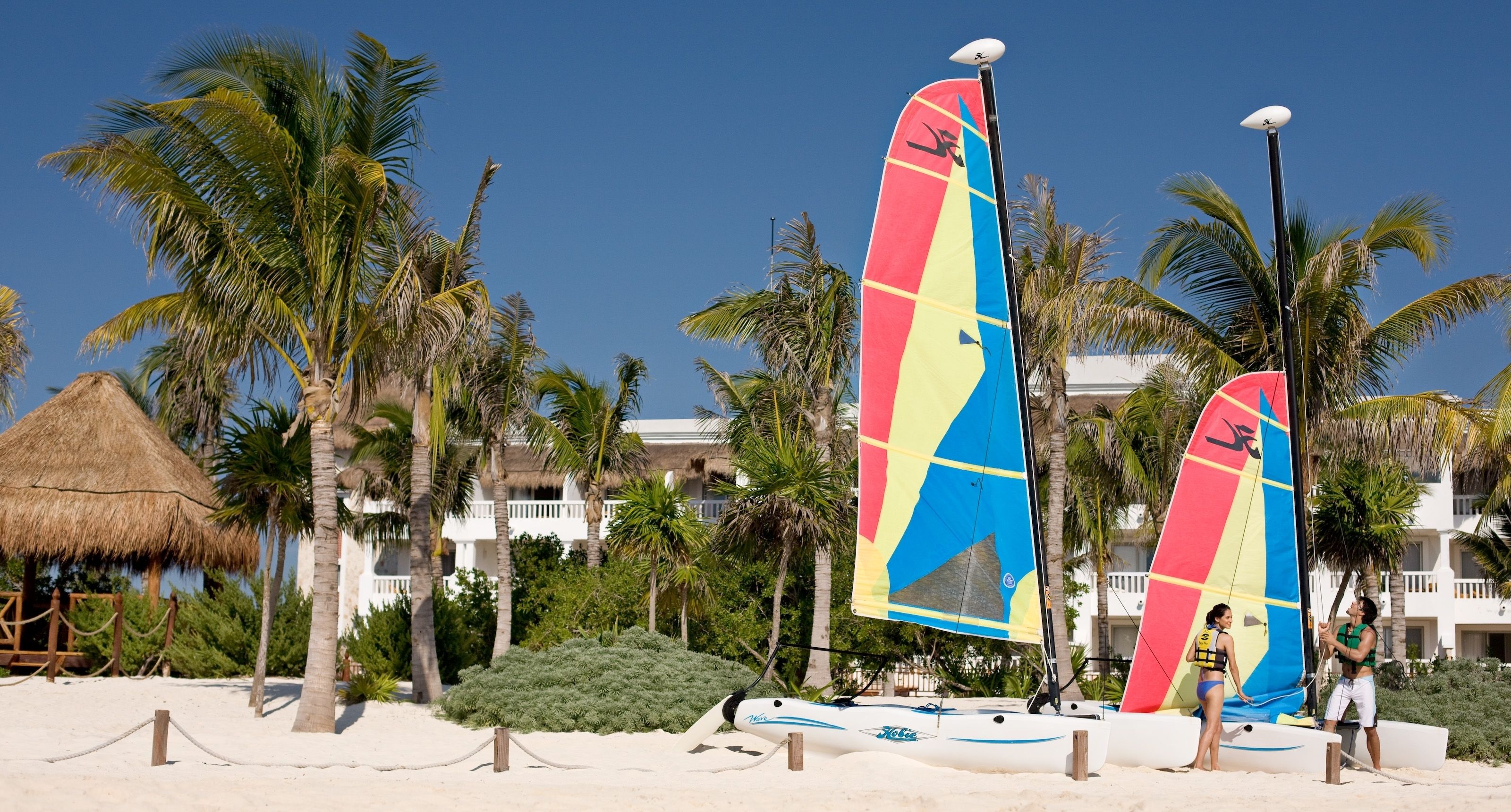 A beach with two sailboats, palm trees, and a thatched-roof structure.