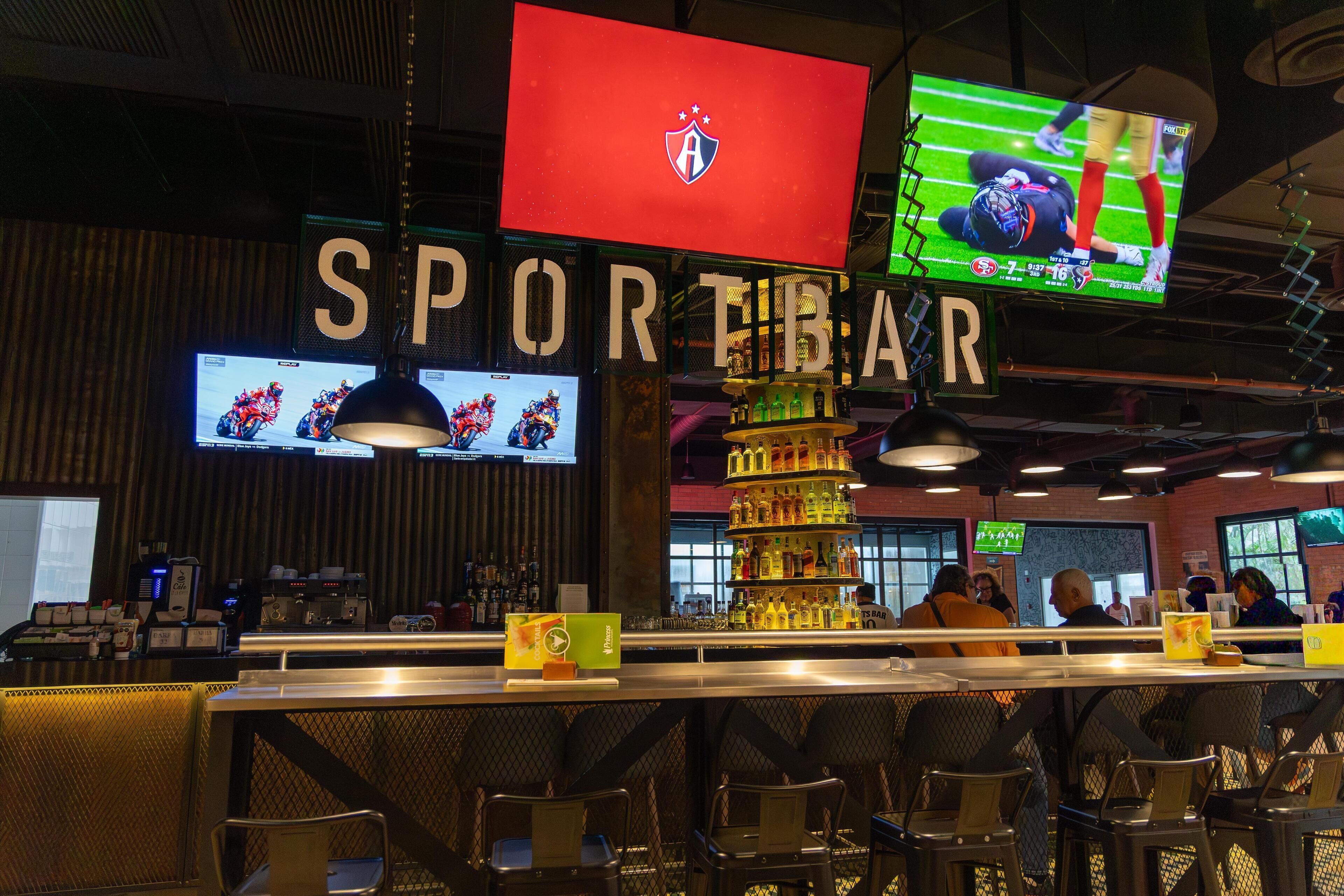 Eine Barbereich mit mehreren Bildschirmen, die Sportinhalte zeigen, einem gut sichtbaren Schild mit der Aufschrift 'SPORT BAR' und einer Auswahl an Getränken in Regalen.