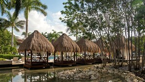 Un resort con cabañas de techo de paja junto a una piscina, rodeado de palmeras y un cielo despejado.