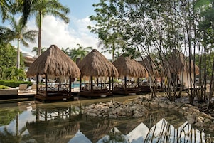 A resort with thatched-roof cabanas by a pool, surrounded by palm trees and a clear sky.