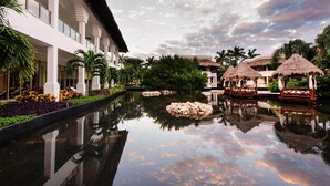 Un resort con piscina, construcciones de techo de paja y un edificio blanco con balcones.