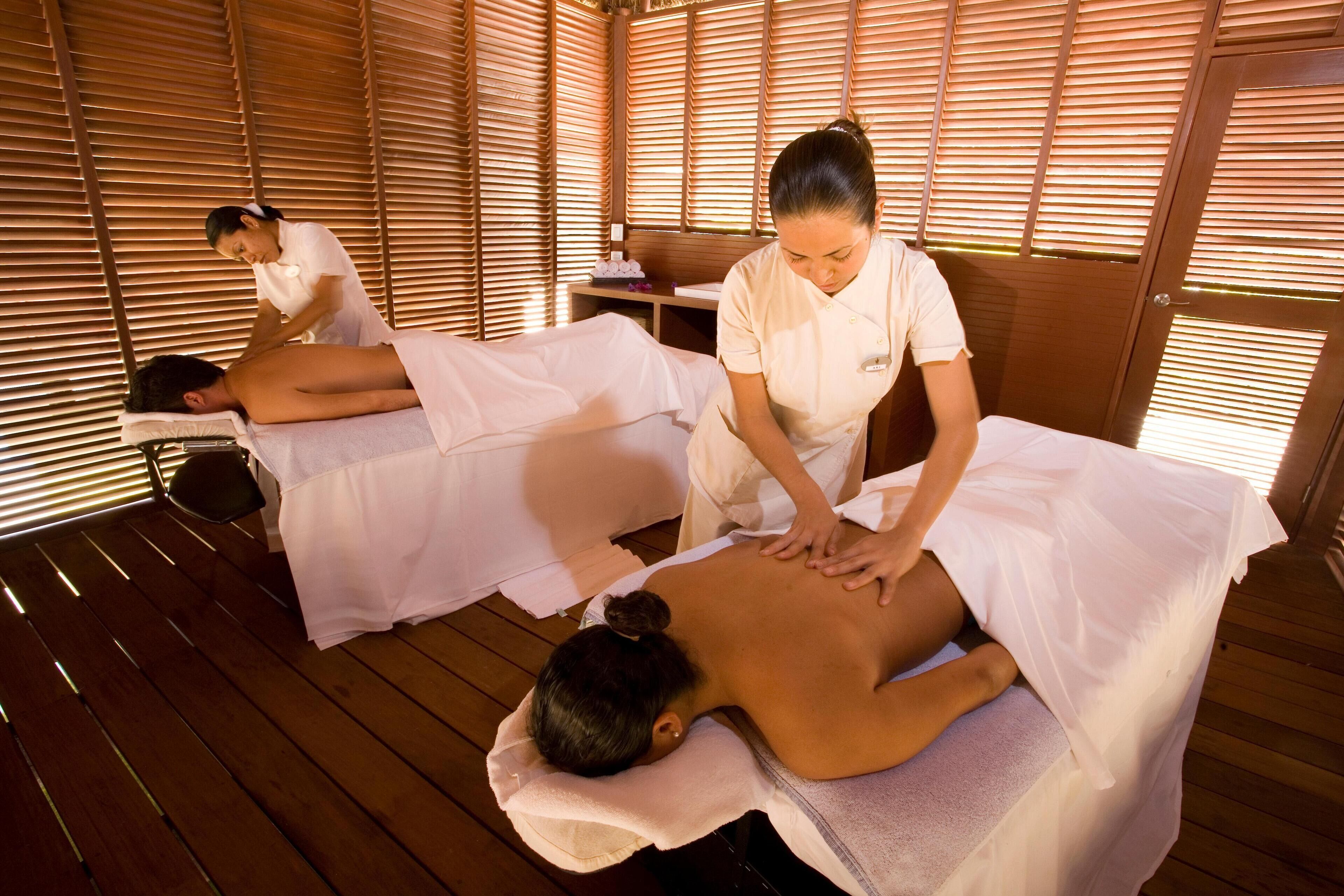Een massageruimte met houten vloeren en wanden, twee massagetafels en een persoon die een rugmassage ontvangt.