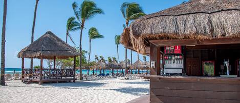 A beach with thatched-roof structures, palm trees, and a Coca-Cola sign.