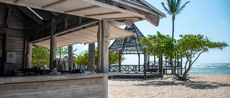 A wooden beach structure with a covered seating area, a palm tree, and a view of the ocean.
