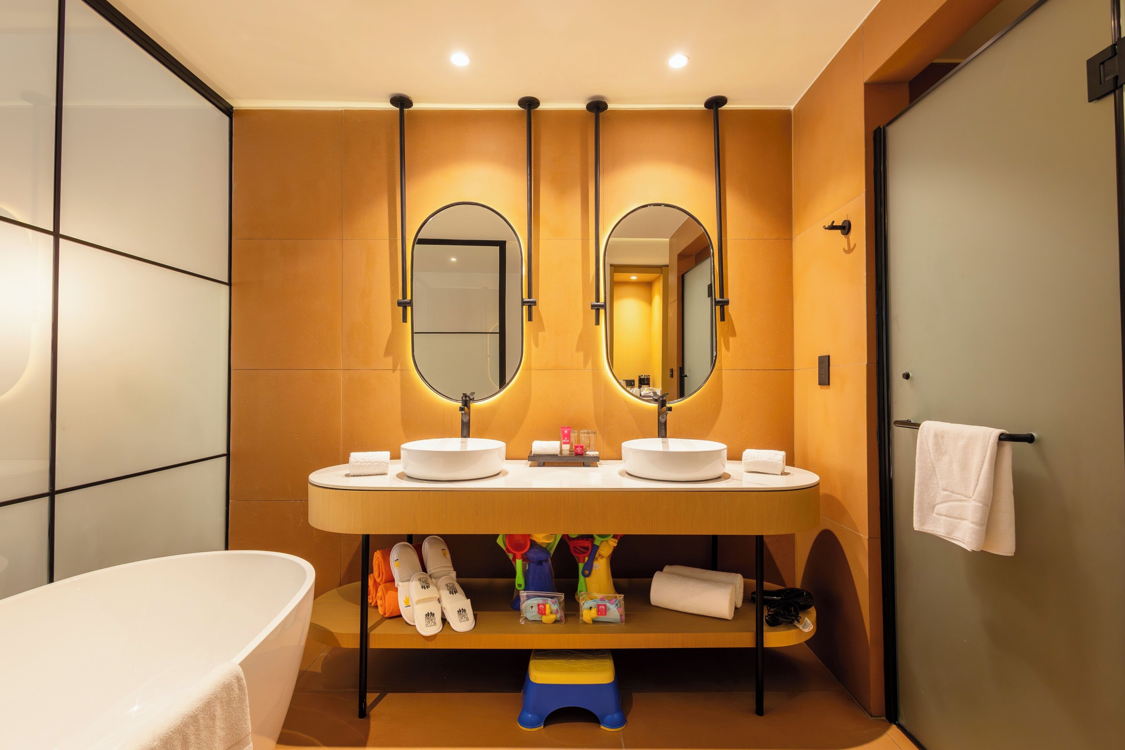 Un baño moderno con bañera, dos lavamanos y un espejo. Hay un estante con artículos de tocador y un taburete.