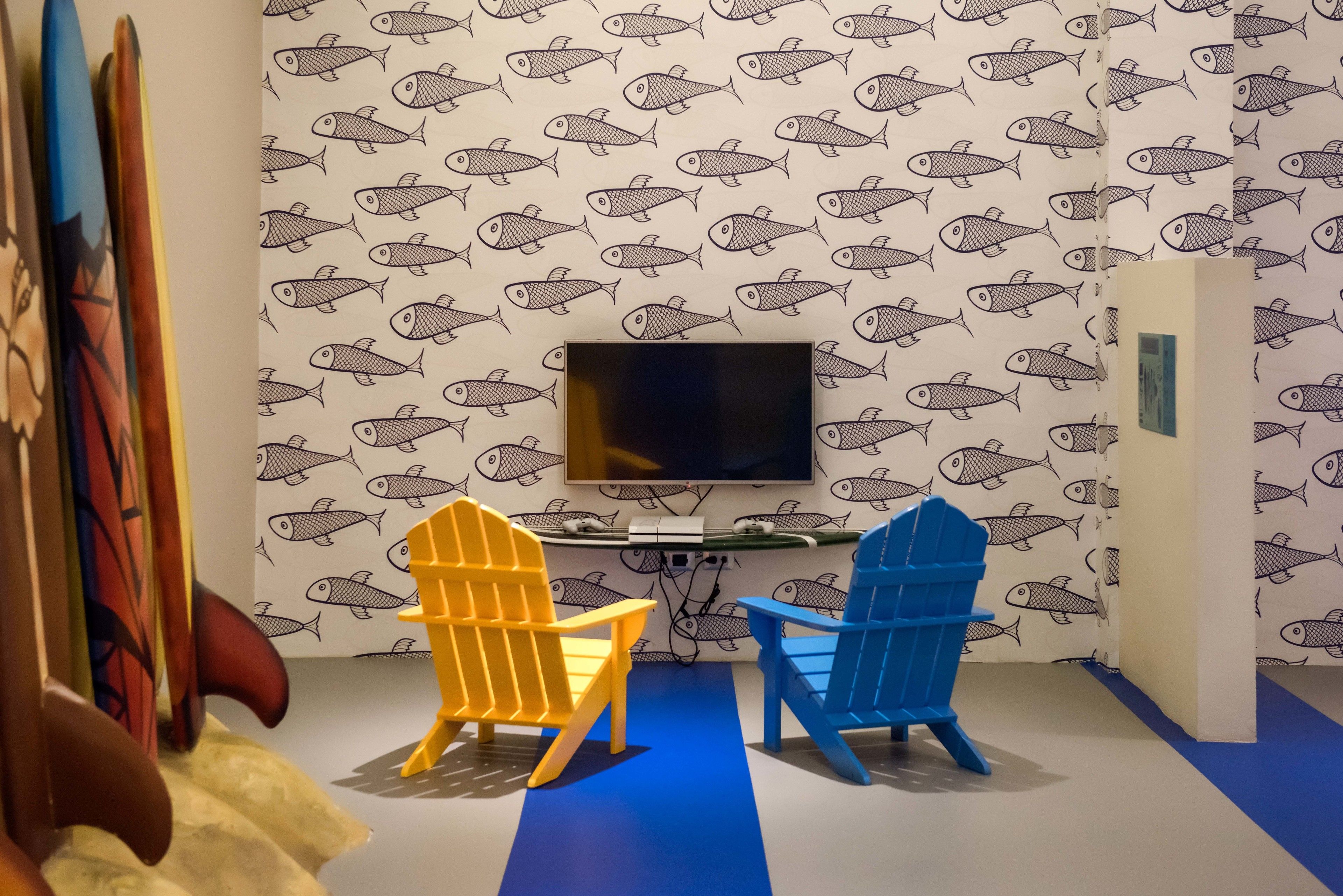 Ein Zimmer mit Fischmuster-Tapete, einem an der Wand befestigten Flachbildfernseher und zwei bunten Adirondack-Stühlen.