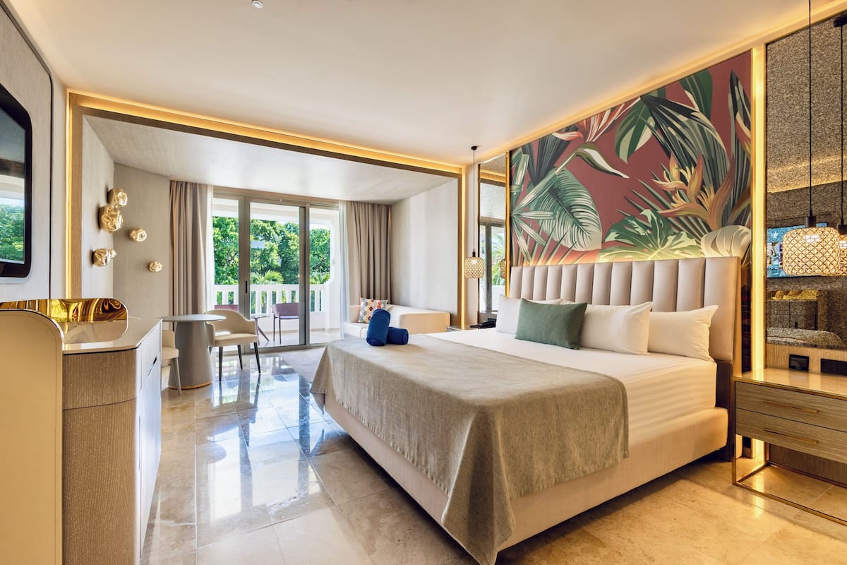 A hotel room with a large bed, a desk, a chair, a balcony with a view, and a tropical-themed wall mural.