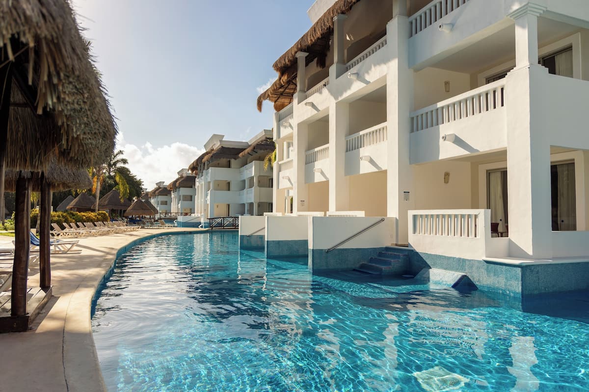 A swimming pool with a clear blue water, surrounded by white buildings with balconies, and a thatched roof structure.