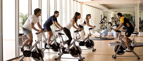 A group of people exercising on stationary bikes in a well-lit indoor space with large windows and a wooden floor.