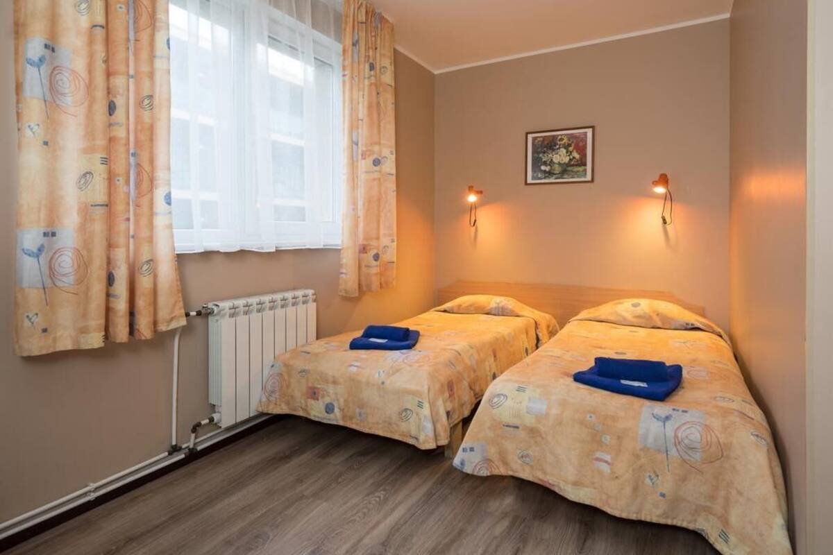 A room with two beds, a radiator, a window with curtains, and wall-mounted lights.