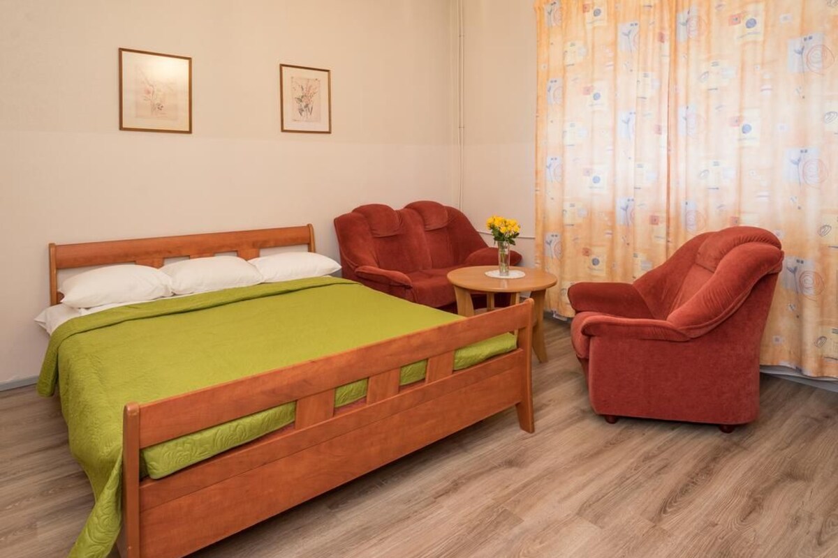 A room with a wooden bed, two red armchairs, a small round table with a vase of flowers, and a curtain with a pattern.