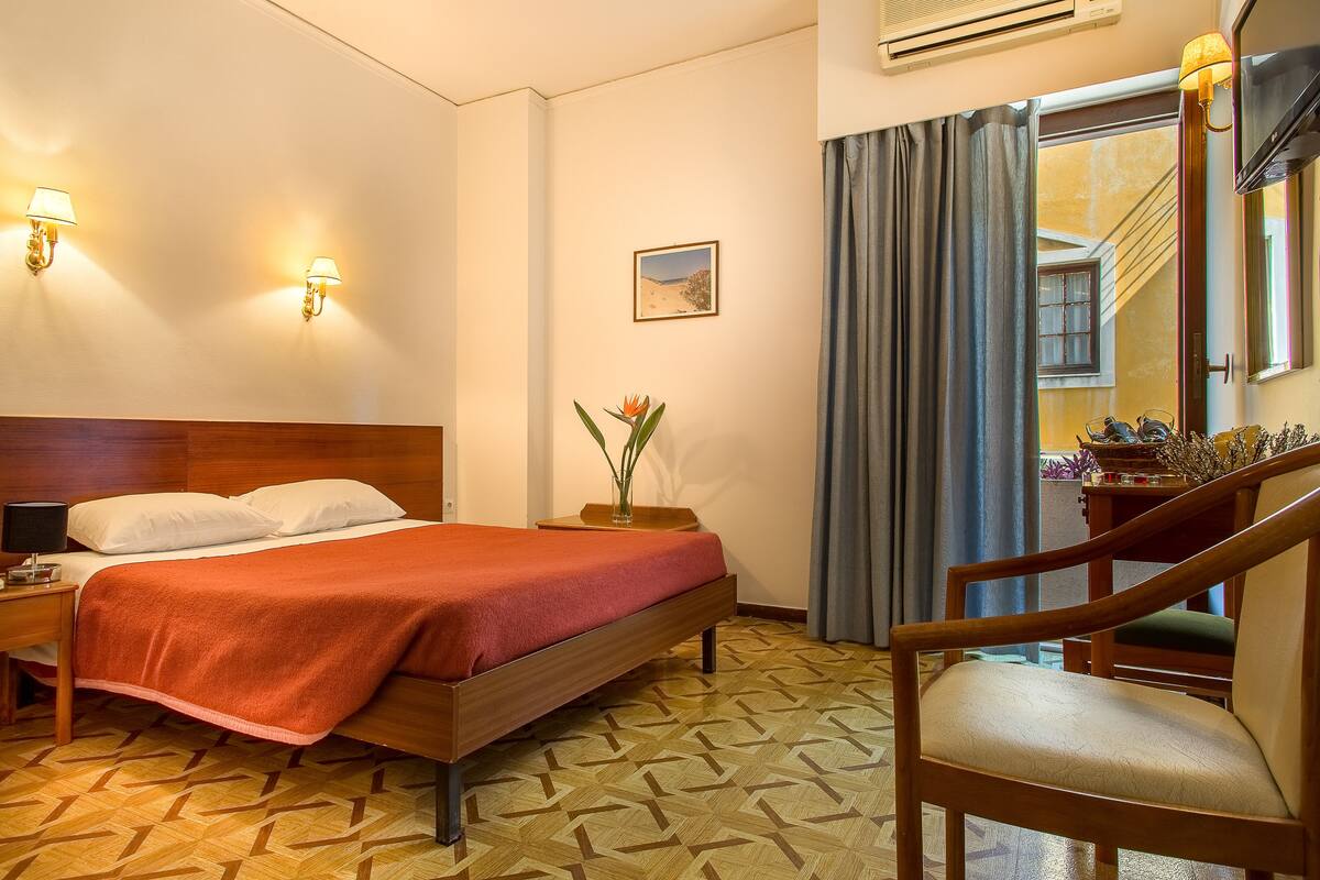 A hotel room with a wooden bed, a nightstand with a lamp, a chair, a small table with a vase, and a picture on the wall.