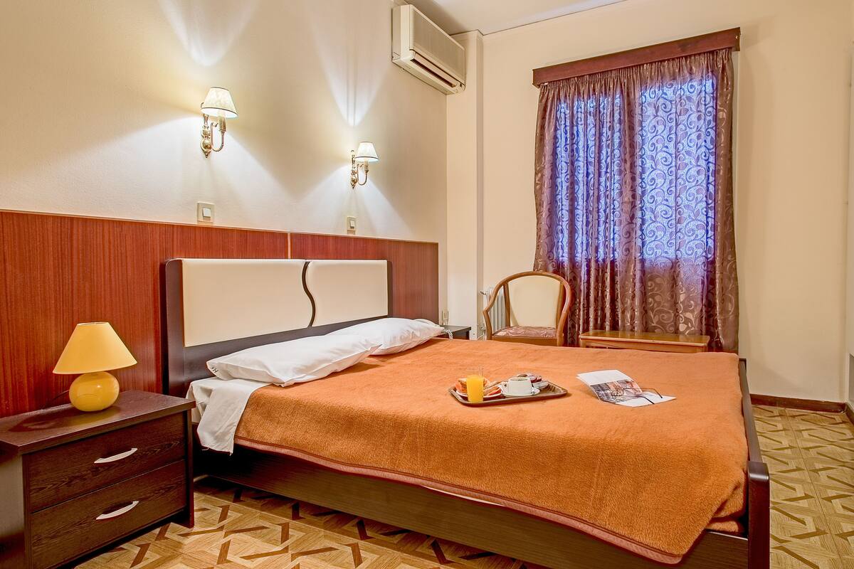 A hotel room with a bed, a nightstand with a lamp, a window with curtains, and a wall-mounted air conditioner.