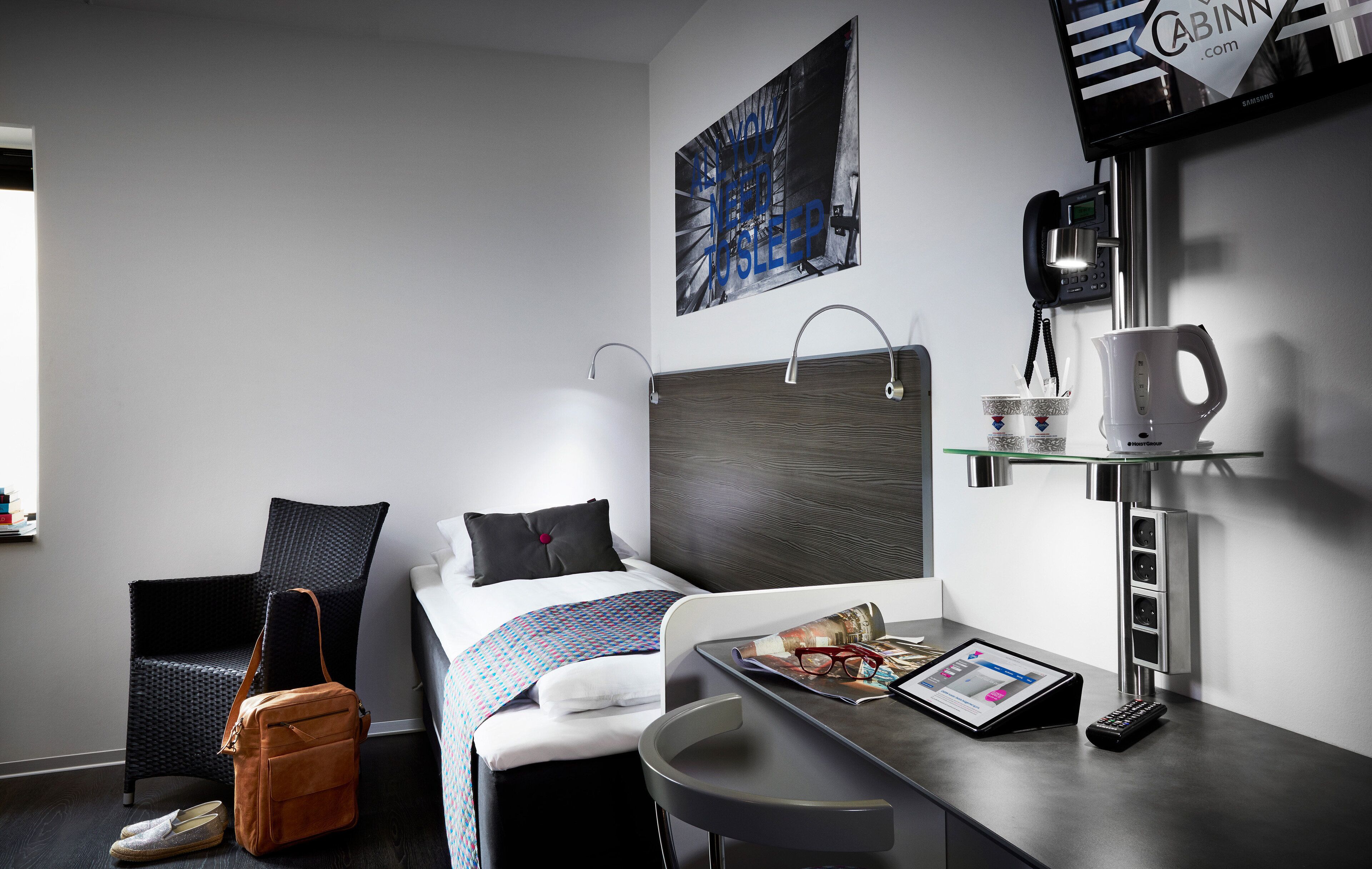 A modern hotel room with a bed, a desk with a tablet, a coffee machine, and a television on the wall.
