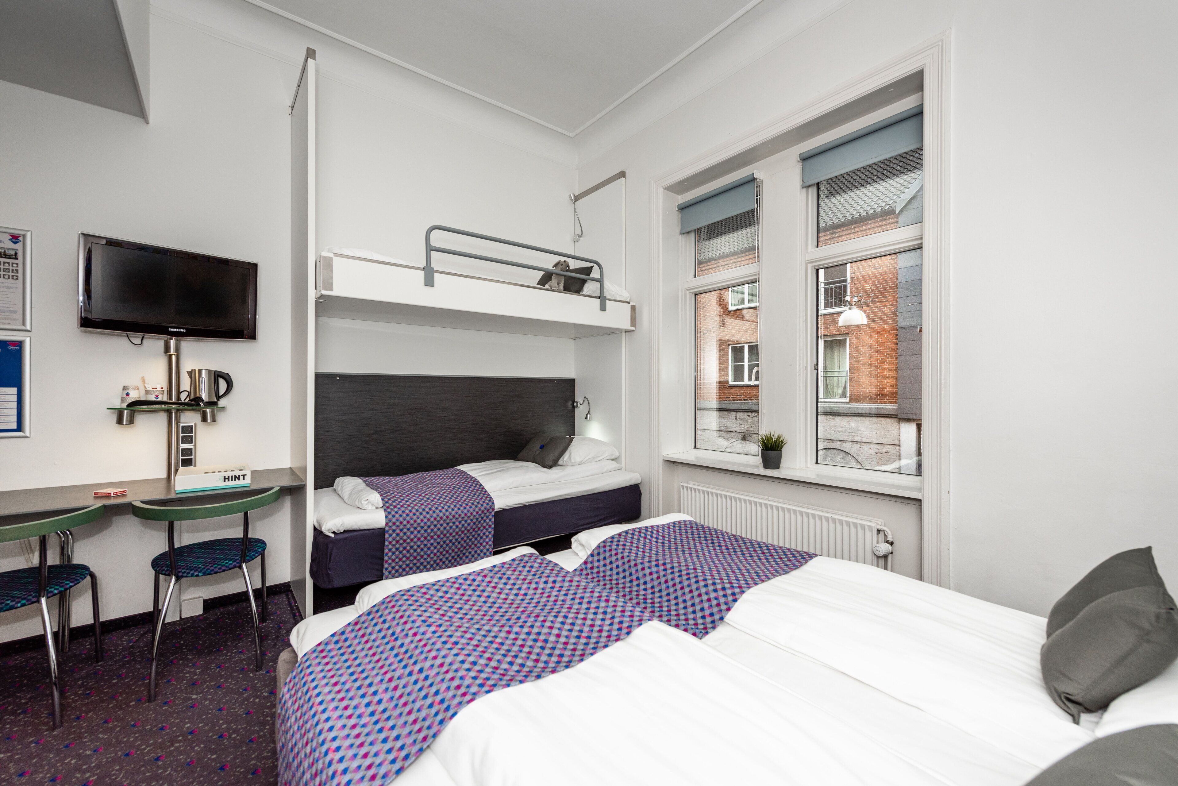 Quarto de hotel com beliches, uma escrivaninha, uma cadeira, uma televisão e janela com vista para o exterior.