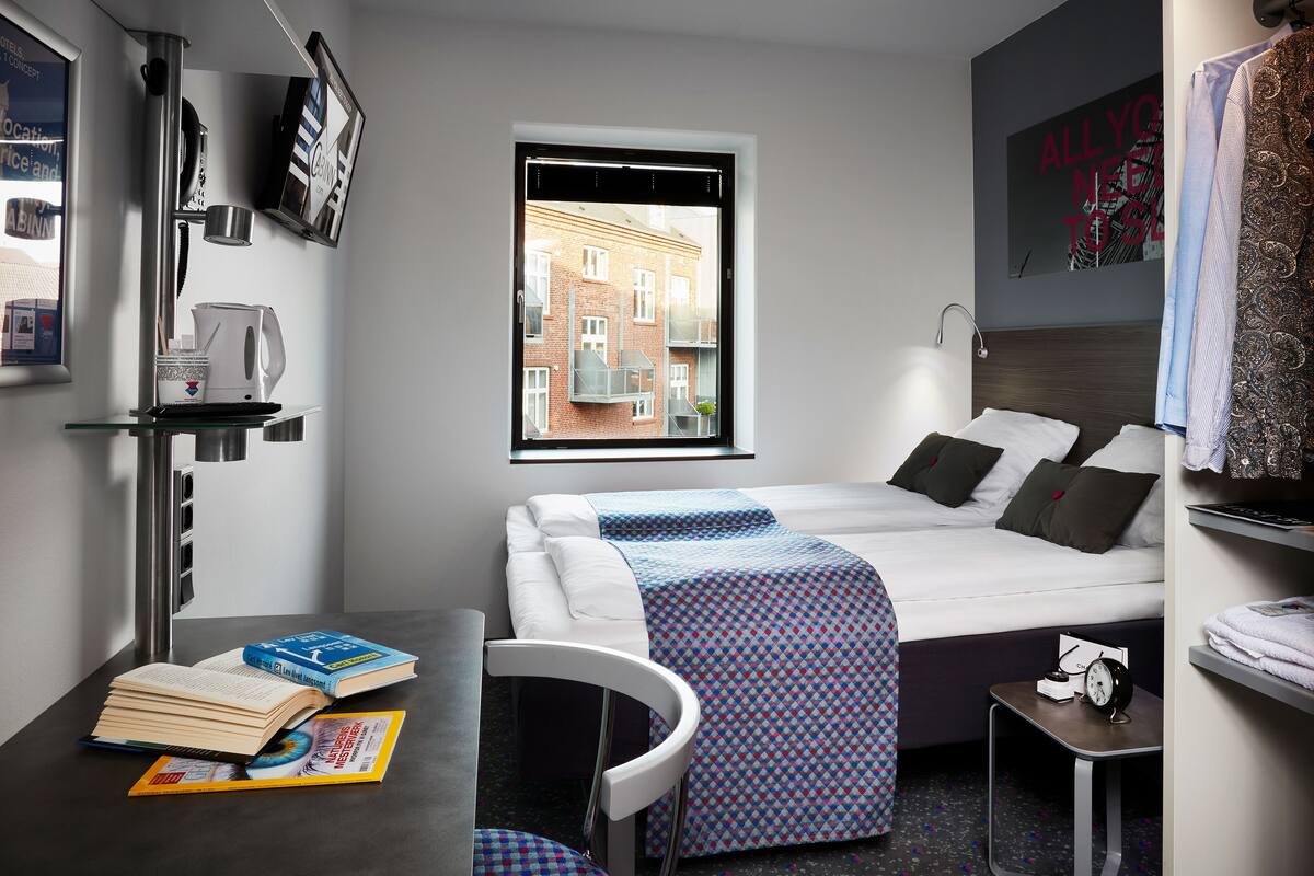 Una habitación de hotel moderna con una cama, un escritorio con libros, una ventana con vista a edificios y una pared con un cartel.