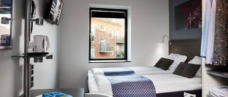 A modern hotel room with a bed, a desk with books, a window with a view of buildings, and a wall with a sign.