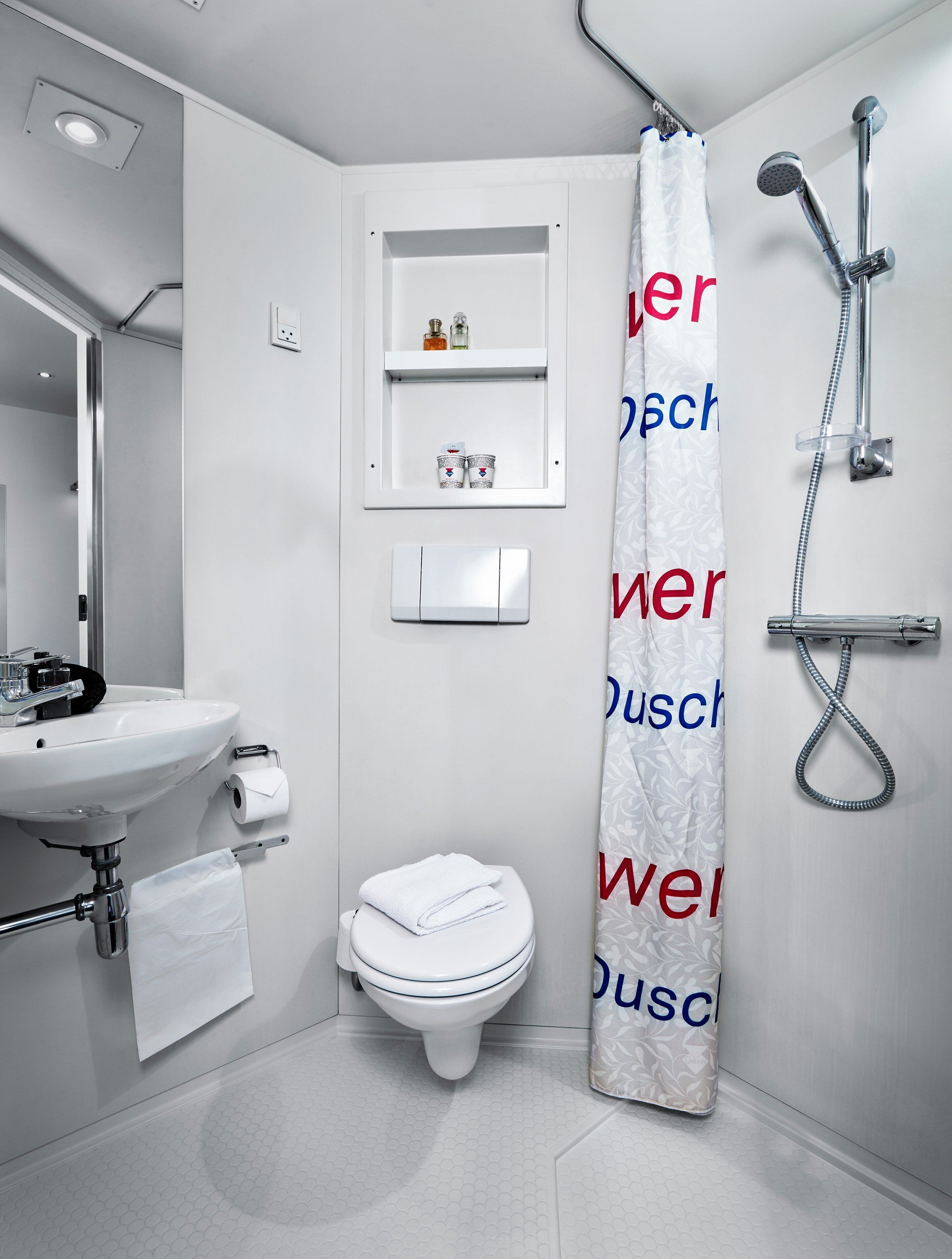 A modern bathroom with a white sink, toilet, and shower. There is a towel rack with a folded towel, a small shelf with toiletries, and a shower curtain with text.