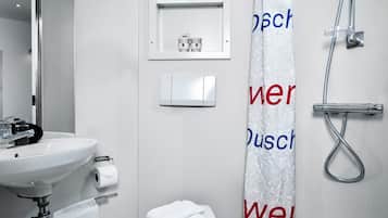 A modern bathroom with a white sink, toilet, and shower. There is a towel rack with a folded towel, a small shelf with toiletries, and a shower curtain with text.