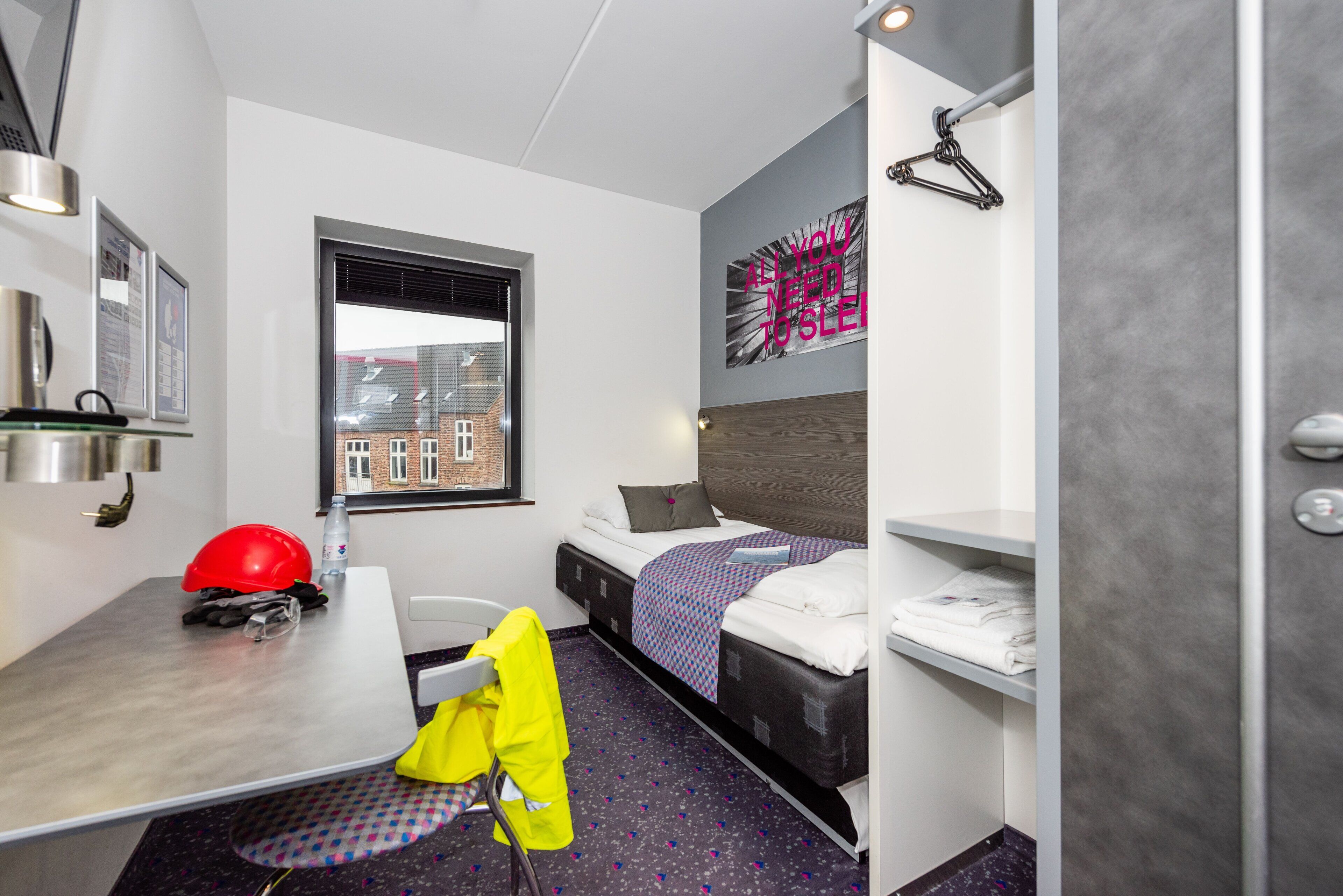 A compact hotel room with a bed, a desk, a chair, a window, and a wall-mounted shelf.
