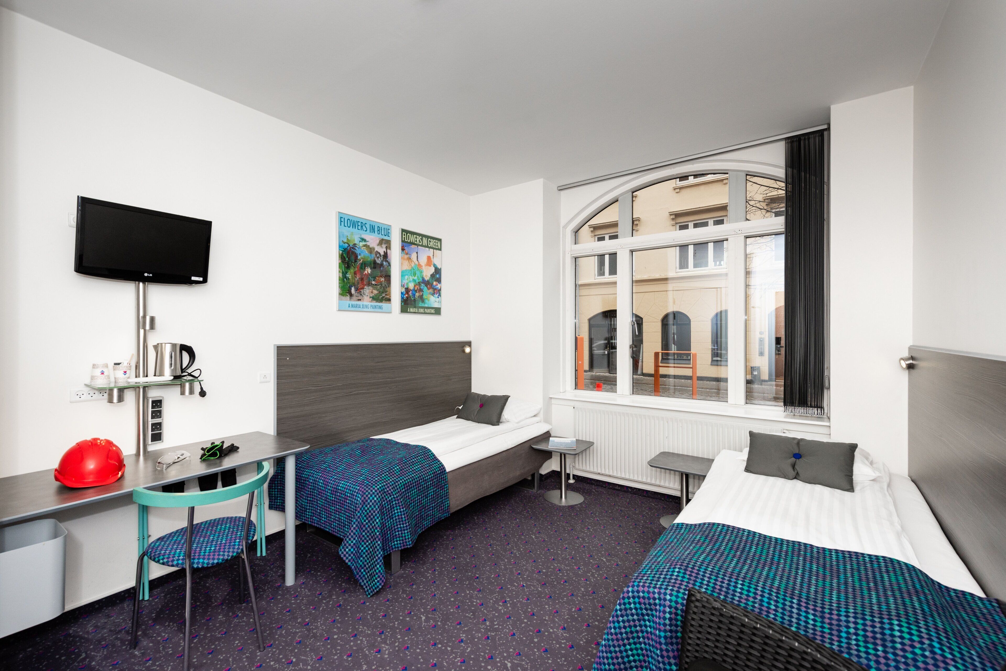 Habitación de hotel con dos camas, un escritorio, una silla, un televisor y una ventana con vistas a edificios.
