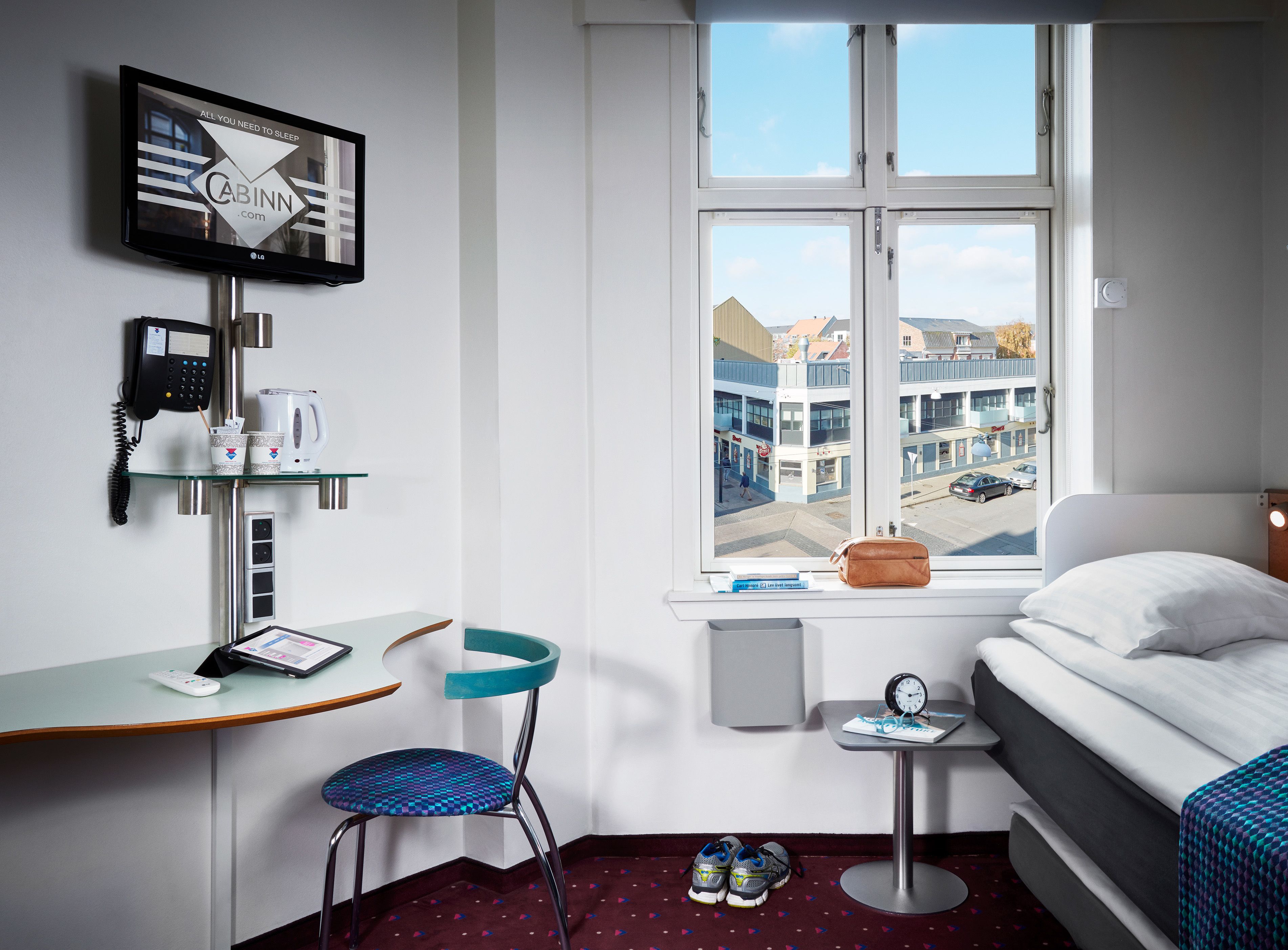 A hotel room with a bed, a desk with a computer, a chair, a window with a view of buildings, and a television on the wall.