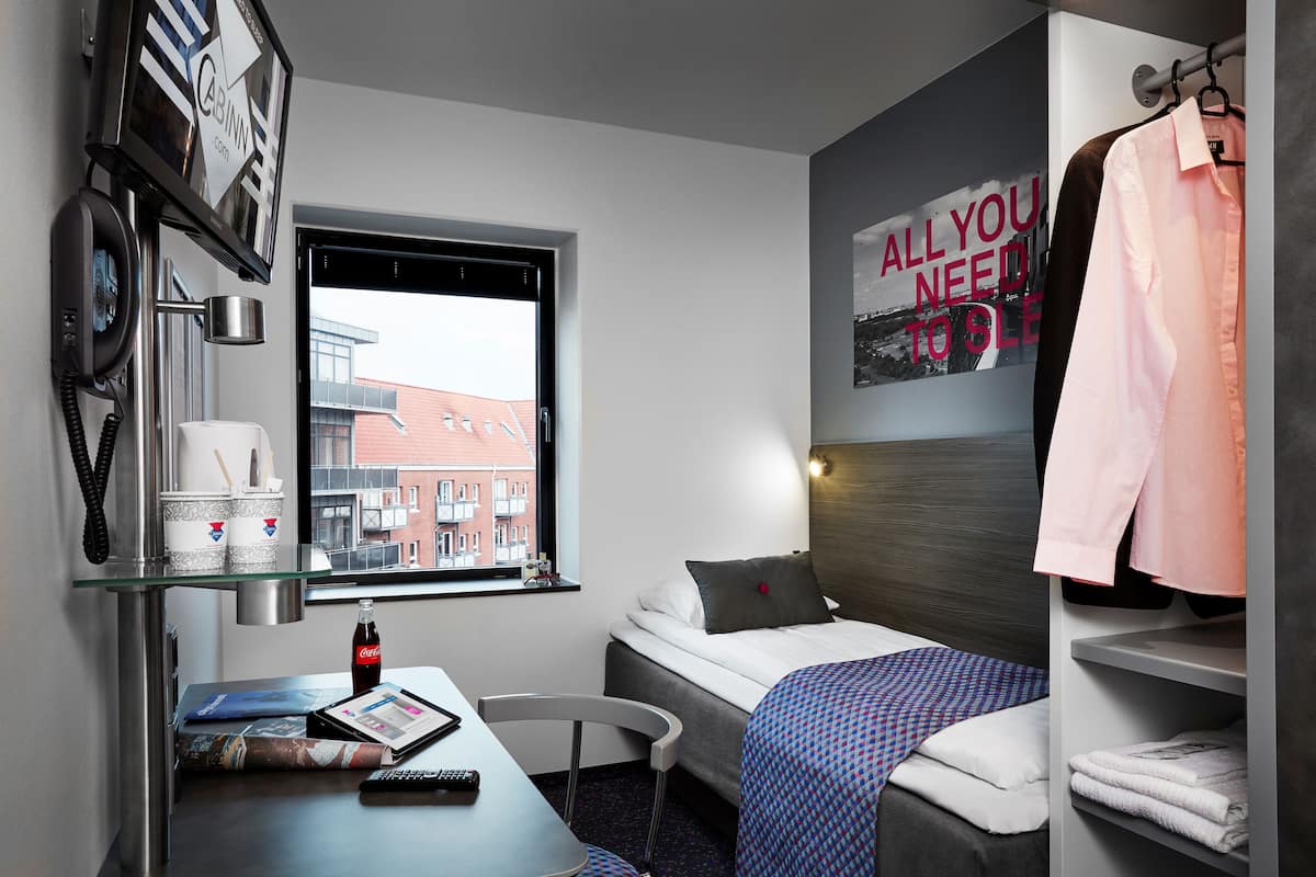 Una habitación de hotel con cama, escritorio con computadora, silla, ventana con vista a edificios y un cartel en la pared.