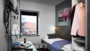 Habitación de hotel con cama, escritorio con computadora, silla, ventana con vista a edificios, perchero con una camisa y un cartel en la pared.