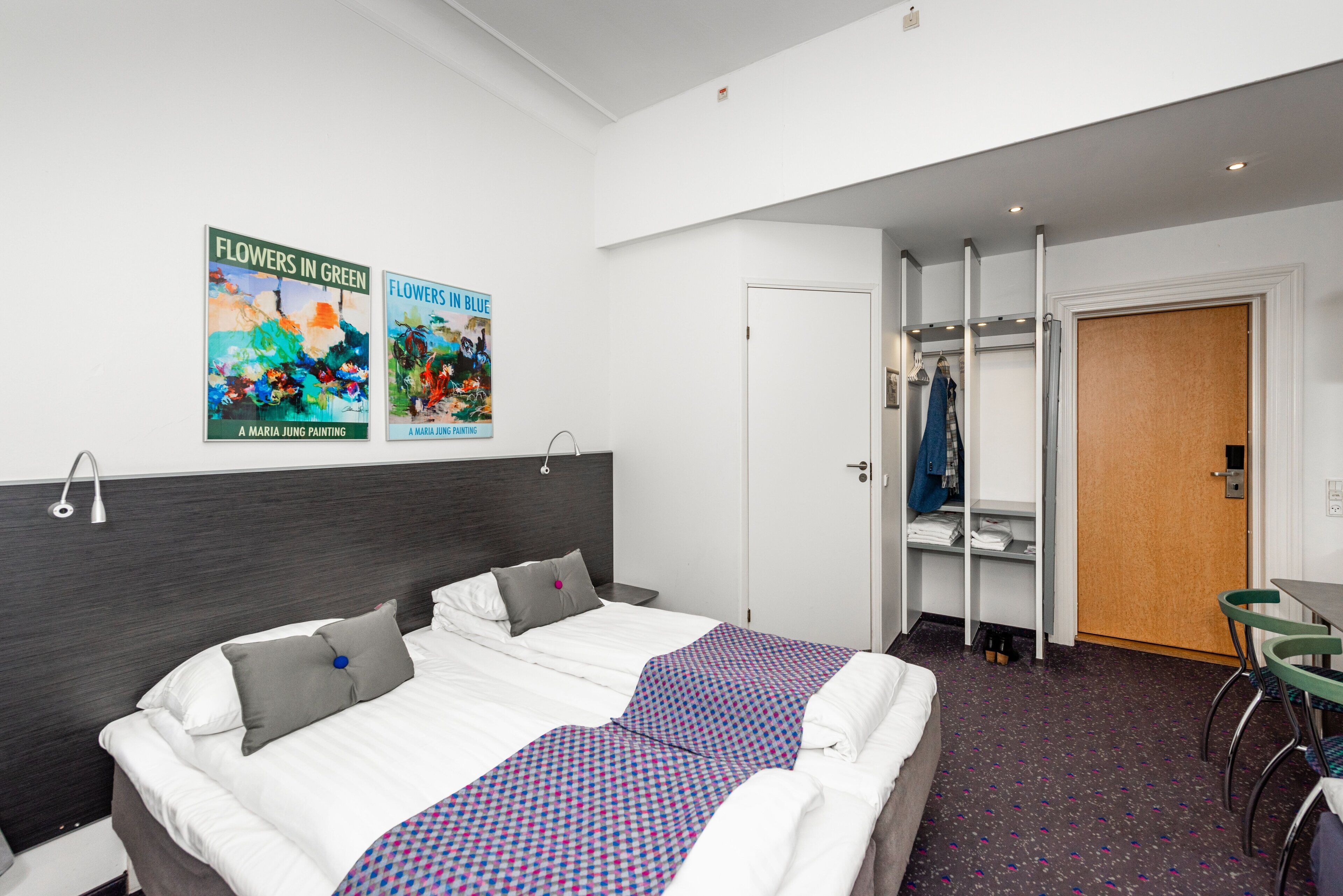 A hotel room with a bed, a wardrobe, a desk, and two paintings on the wall.