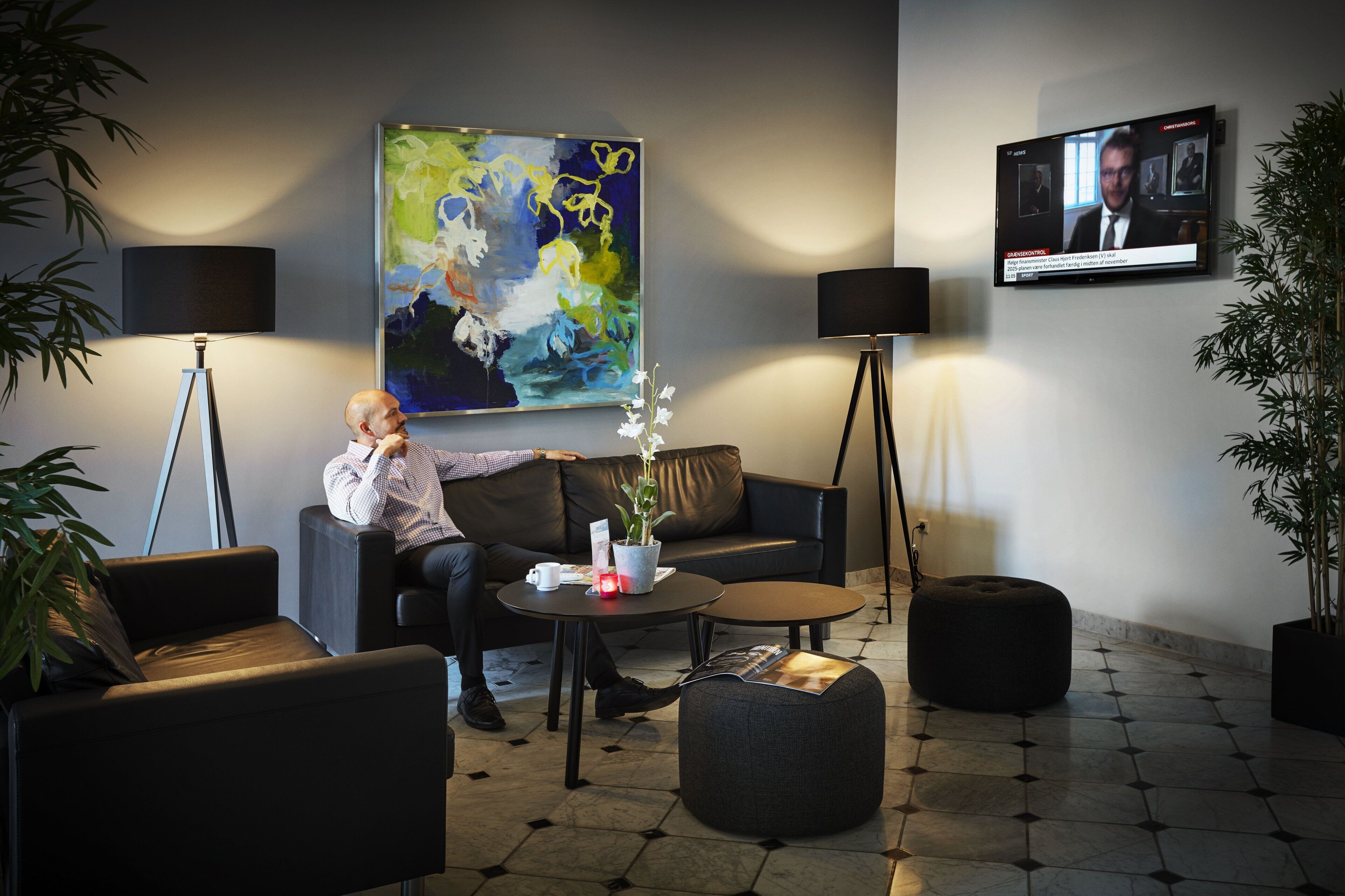 Een moderne lobby met een bank, fauteuils, een bijzettafeltje, een potplant, een schilderij en een televisiescherm.