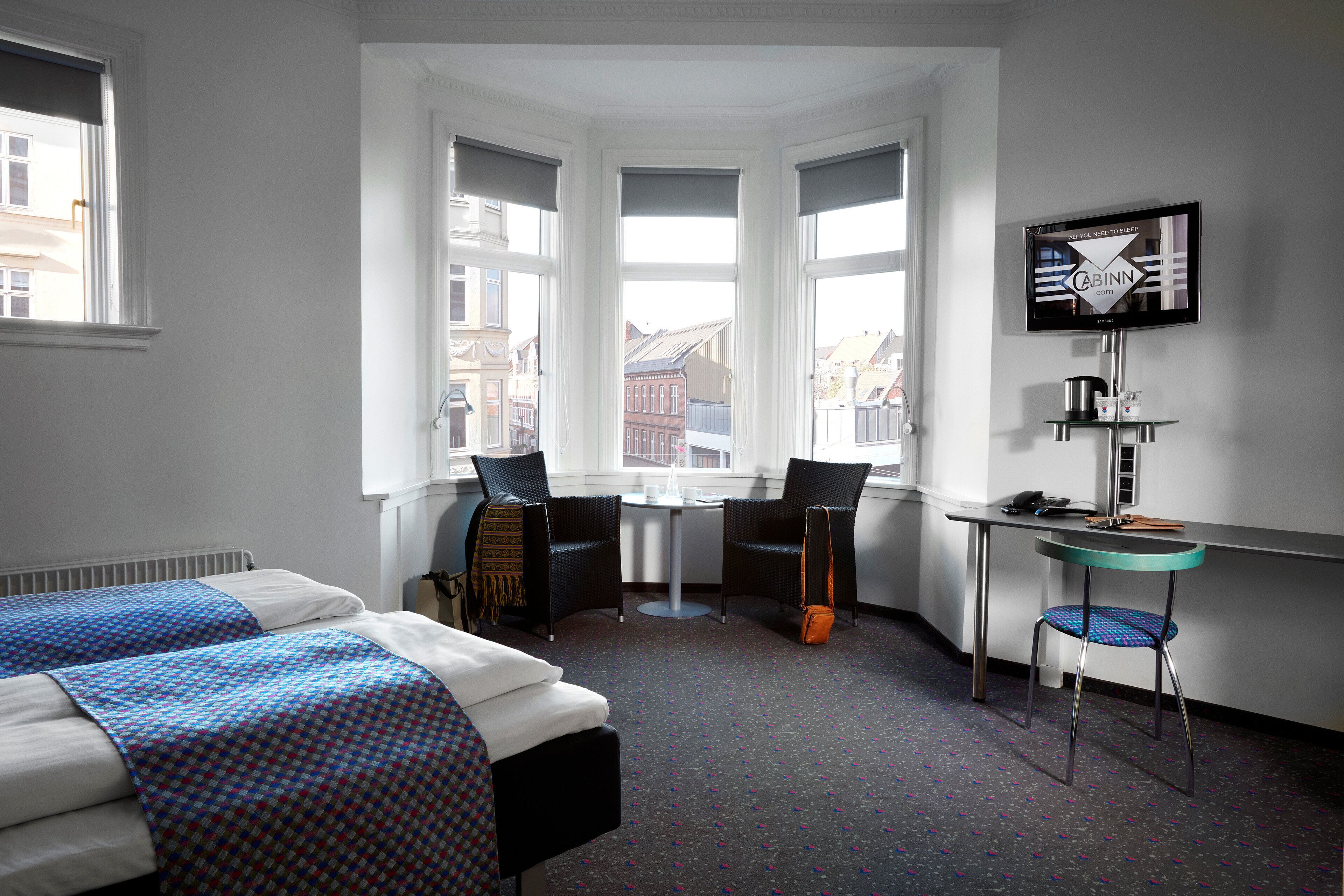 Habitación de hotel con dos camas, un escritorio, una silla, un televisor y vistas a edificios a través de amplios ventanales.