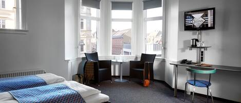 Habitación de hotel con dos camas, un escritorio, una silla, un televisor y vistas a edificios a través de amplios ventanales.