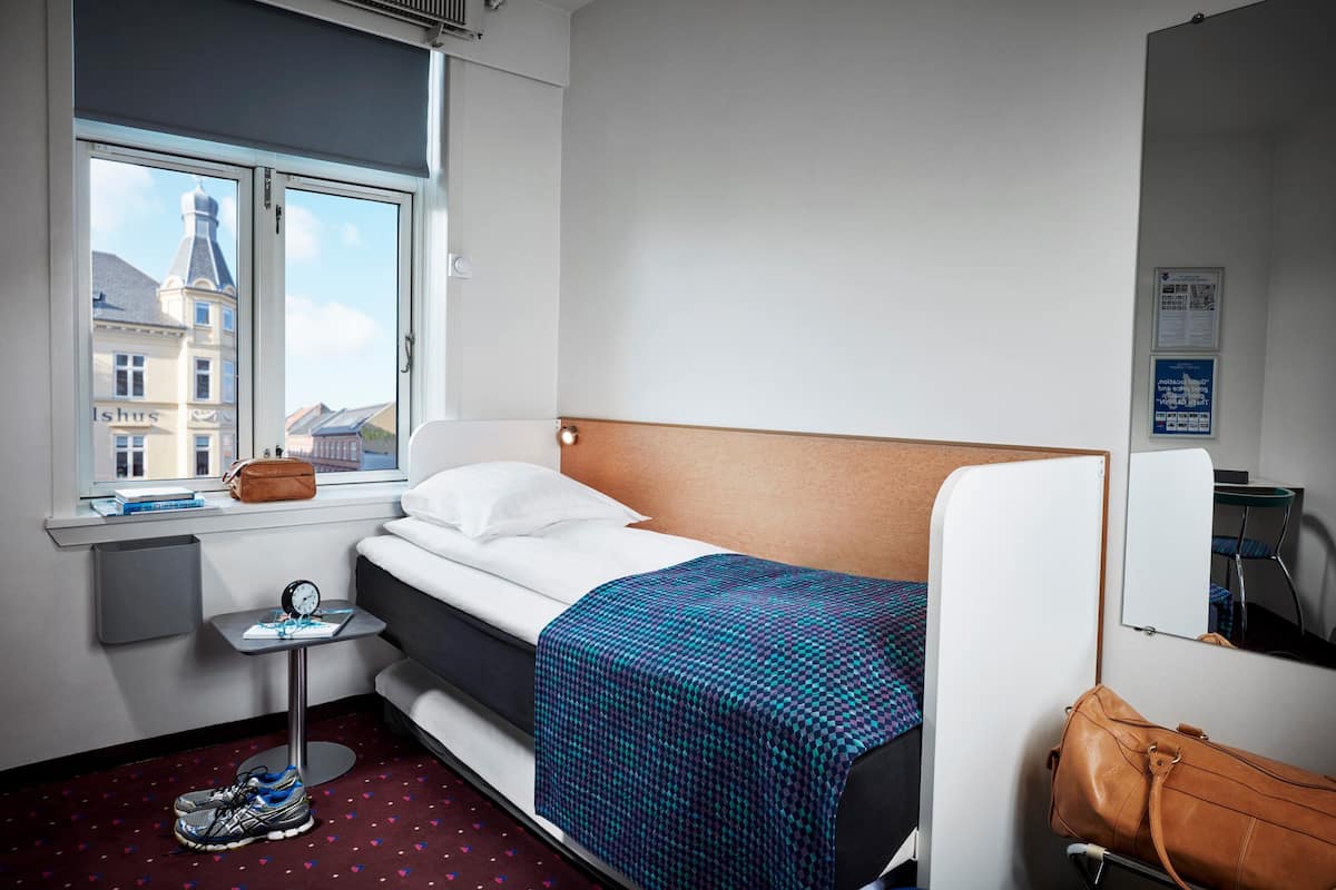 Habitación de hotel con cama, una mesita de noche con un reloj, una ventana con vistas a edificios y una maleta en el suelo.